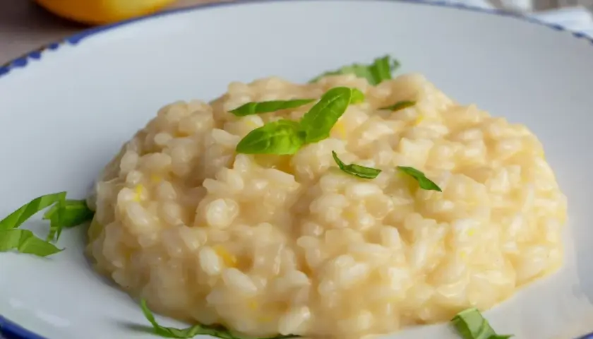 Kremowe risotto cytrynowe, ozdobione listkami bazylii, na talerzu z cytryną w tle.