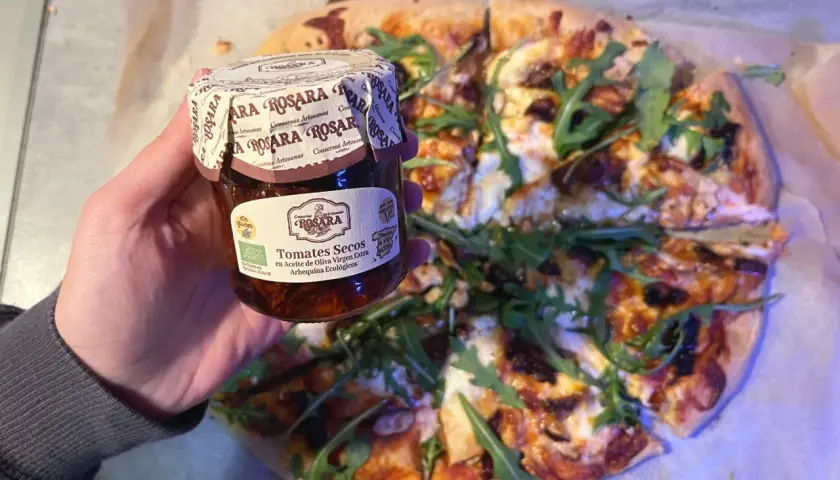 Mano sostiene un tarro de tomates secos en aceite de oliva virgen extra, listo para añadir a una pizza con rúcula.