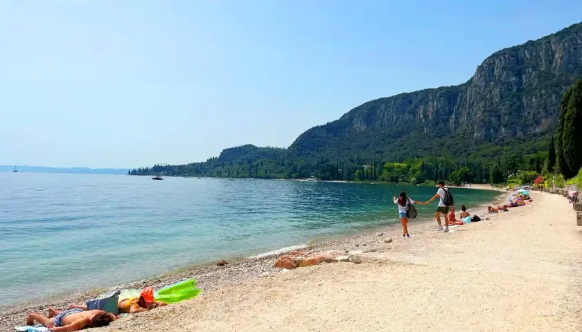 Ein Paar hält Händchen am Strand, während andere am Gardasee schwimmen. Berge und Bäume säumen die Küste.