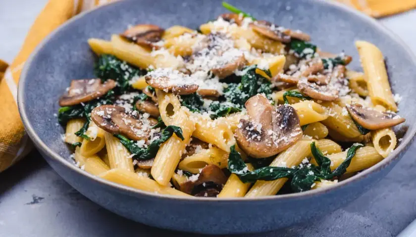 Penne z pieczarkami i szpinakiem, posypane parmezanem, podane w szarej misce.