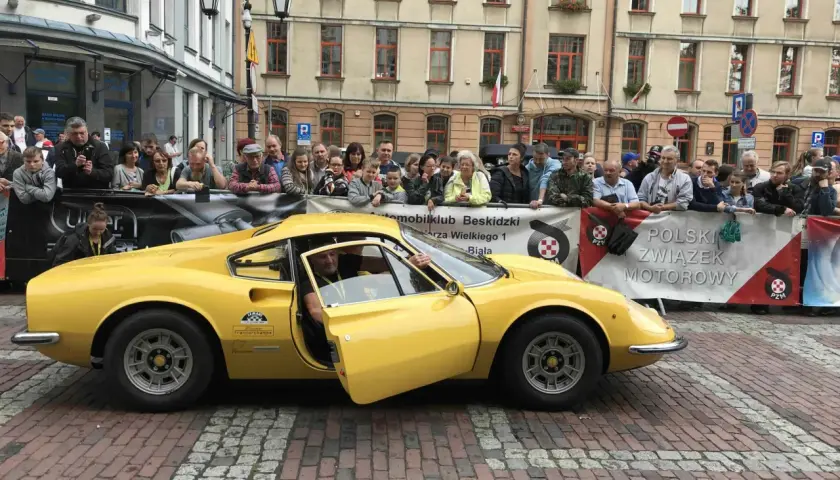 Żółte Ferrari Dino z otwartymi drzwiami, kierowca w środku, tłum widzów za barierką.
