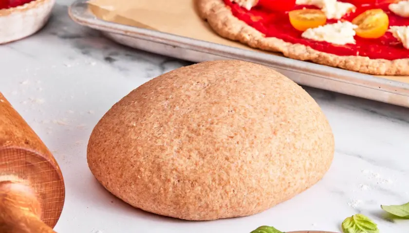 Ein runder Laib vollkorn pizzateig liegt bereit. Im Hintergrund eine fast fertige Pizza mit Tomaten und Mozzarella.