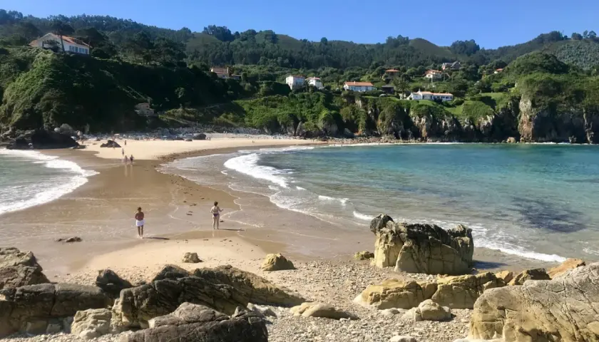 La **playa de Amió** es un paraíso de arena dorada y aguas cristalinas, con colinas verdes y casas blancas al fondo.