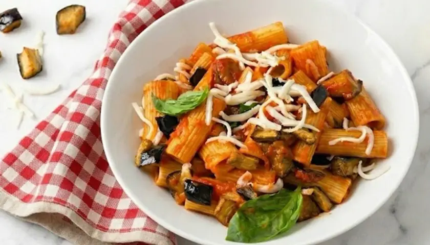 Un plato de pasta alla norma con berenjenas fritas, salsa de tomate y queso rallado, decorado con albahaca fresca.