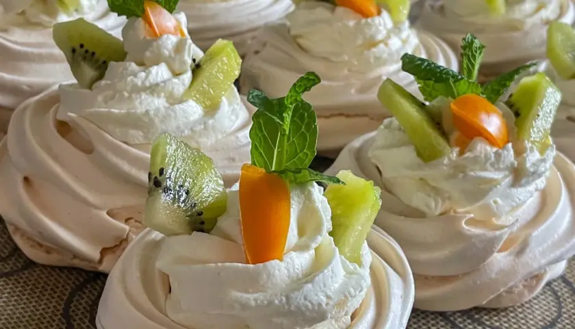 Pyszne gniazdka bezowe z kremem mascarpone, ozdobione kawałkami kiwi, pomarańczy i listkami mięty.
