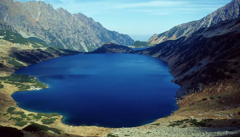 Głębokie, granatowe jezioro otoczone skalistymi zboczami i trawami. Widok na piękne doliny w Tatrach, gdzie natura ukazuje swoją surową, ale zachwycającą twarz.