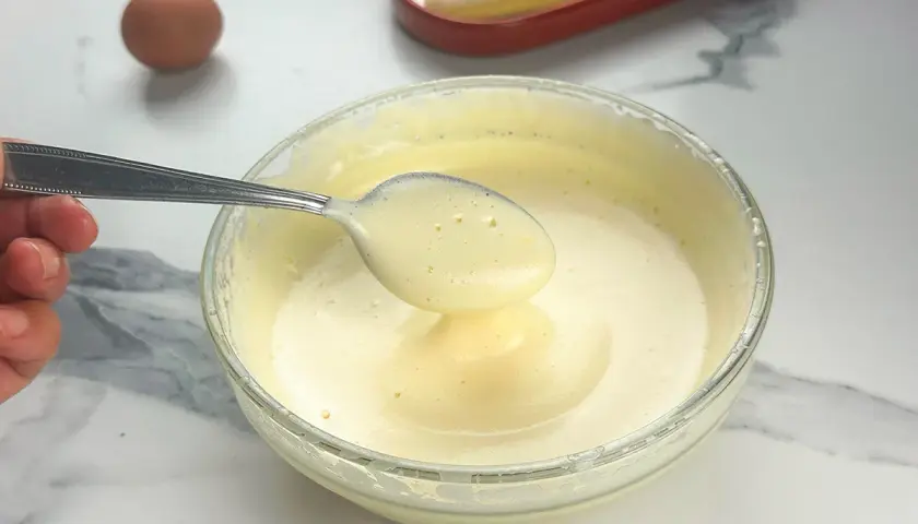 Préparation d'un tiramisu : une cuillère de pâte à bombe aérienne est prélevée d'un bol en verre. Un œuf et des biscuits à la cuillère sont visibles.