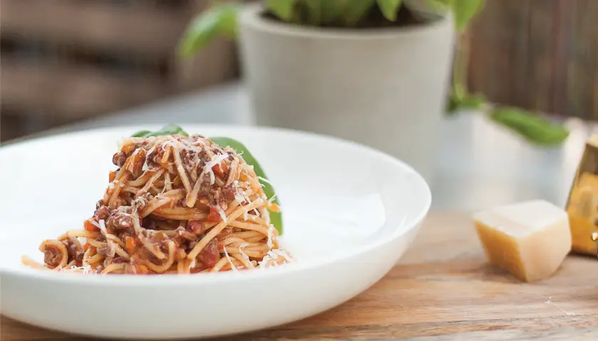 Pyszne spaghetti z bogatym sosem bolońskim, posypane parmezanem, podane na białym talerzu. Obok kawałek sera i listek bazylii.