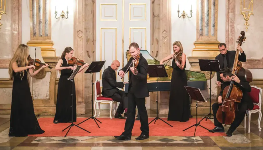 Ensemblen ger en klassisk musik konsert i en elegant sal. Sex musiker spelar fioler, cello och cembalo.