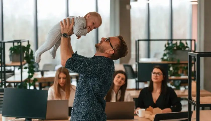 Mężczyzna w okularach podnosi dziecko do góry w biurze, w tle pracują kobiety.