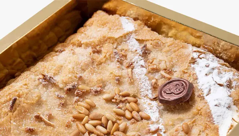 Deliciosa coca de Llavaneres con piñones y un toque de azúcar glas, presentada en una caja dorada.