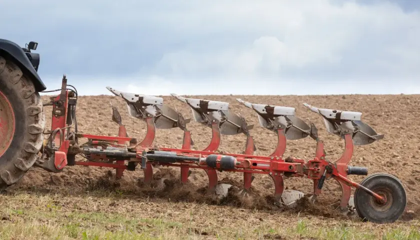Czerwony pług z wieloma skibami pracuje na polu, przygotowując ziemię do siewu.