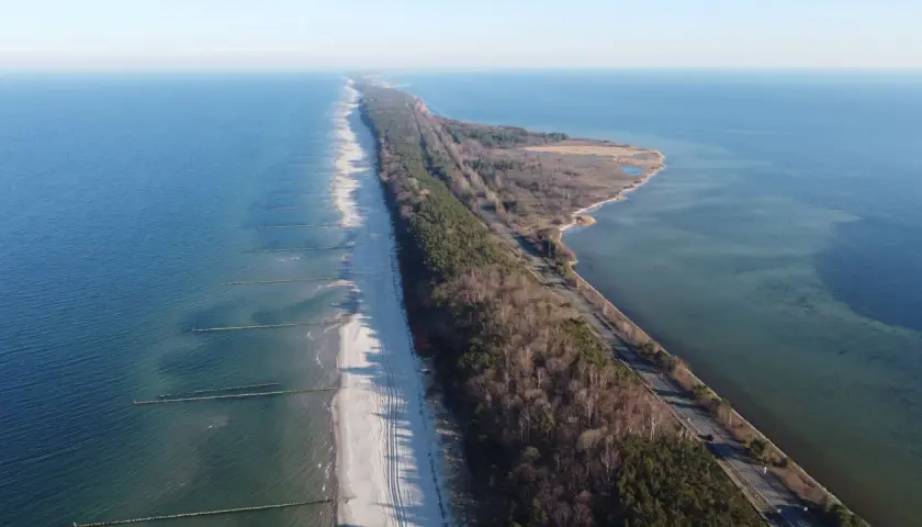 Wąski pas lądu, czyli **półwysep helski**, otoczony przez morze i zatokę. Na plaży widać falochrony, a w głębi las i drogę.