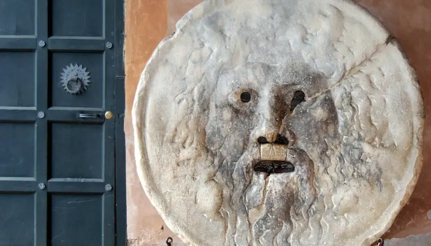 La Bocca della Verità, un volto scolpito nella pietra, sembra pronto a giudicare chiunque osi mentire. Accanto, una porta scura.