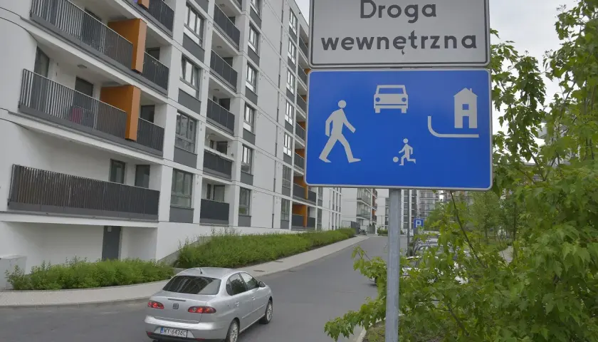 Znak "Droga wewnętrzna" informuje o strefie zamieszkania. Widać tu pieszych, samochód i dom.