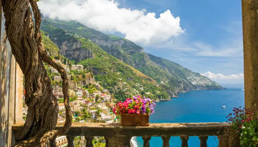 A vibrant view of Amalfi Coast towns clinging to steep green hillsides, overlooking the sparkling blue sea. A twisted tree frames the scene, with a planter of colorful flowers on the balcony railing.