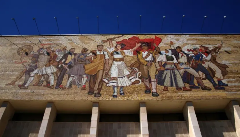 Socrealizm co to? Wielki mozaikowy mural przedstawiający ludzi w strojach ludowych i wojskowych, z flagami i bronią, symbolizujący walkę i jedność.
