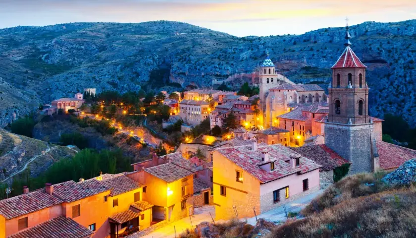 Pueblo español iluminado al anochecer, ideal para escapadas fin de semana España. Casas con tejados de teja y torres de iglesias se alzan entre colinas.