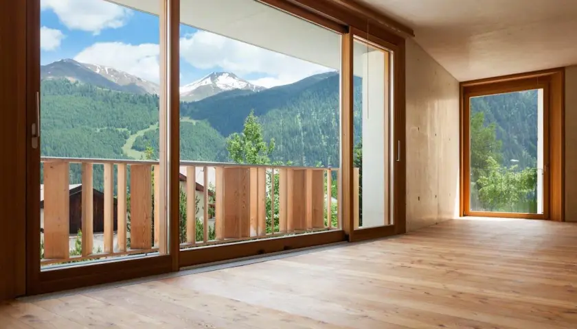 Interno con pavimento in legno e vista panoramica sulle montagne. L'arredamento con infissi marroni crea un'atmosfera calda e accogliente.