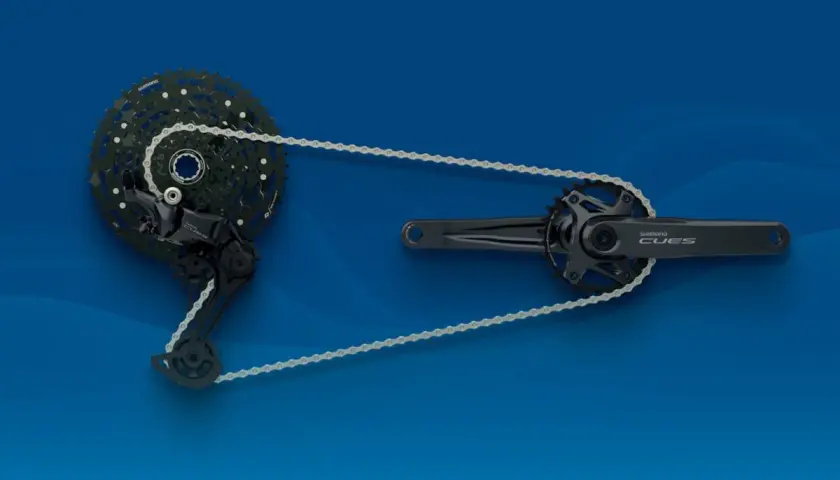 Napęd rowerowy Shimano CUES: korba, przerzutka i kaseta połączone łańcuchem na niebieskim tle.