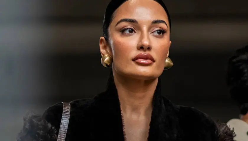 Mujer con peinado pulido y maquillaje natural, luciendo aretes dorados y un abrigo negro de pelo. Su **clean look** irradia elegancia.