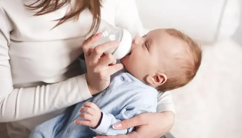 Mama karmi butelką noworodka. Dziecko w niebieskim pajacyku pije mleko, a mama trzyma je czule.