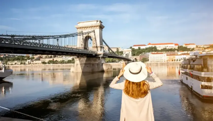 Kobieta w kapeluszu podziwia Most Łańcuchowy w Budapeszcie. W tle widok na Dunaj i budynki, a w portfelu czeka budapeszt waluta na dalsze zwiedzanie.