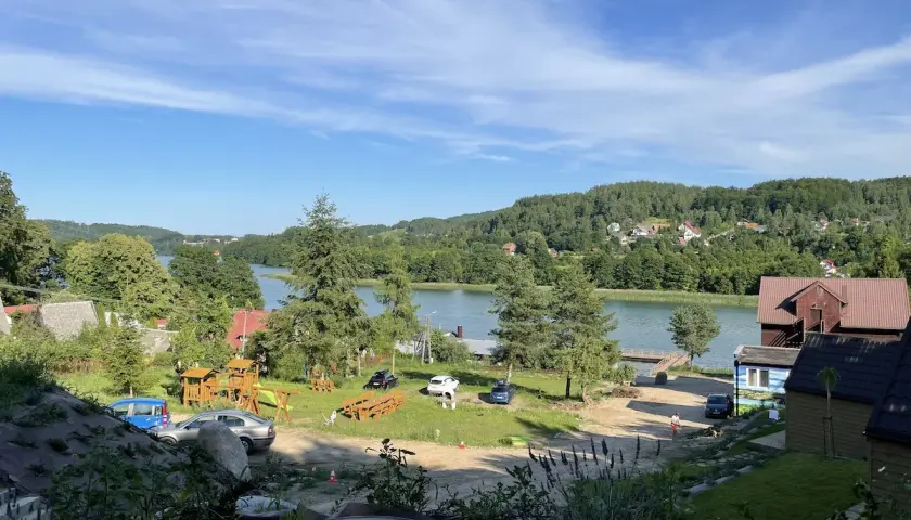 Widok na jezioro i ostrzycki las. Na pierwszym planie plac zabaw i samochody, w tle domki i zielone wzgórza.