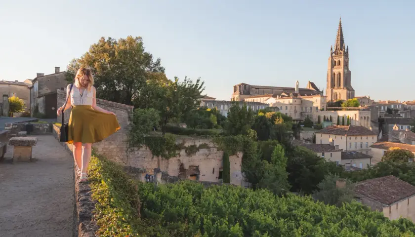 Une femme en jupe jaune marche sur un muret, surplombant le village de Saint-Émilion. La route des vins s'étend à perte de vue.