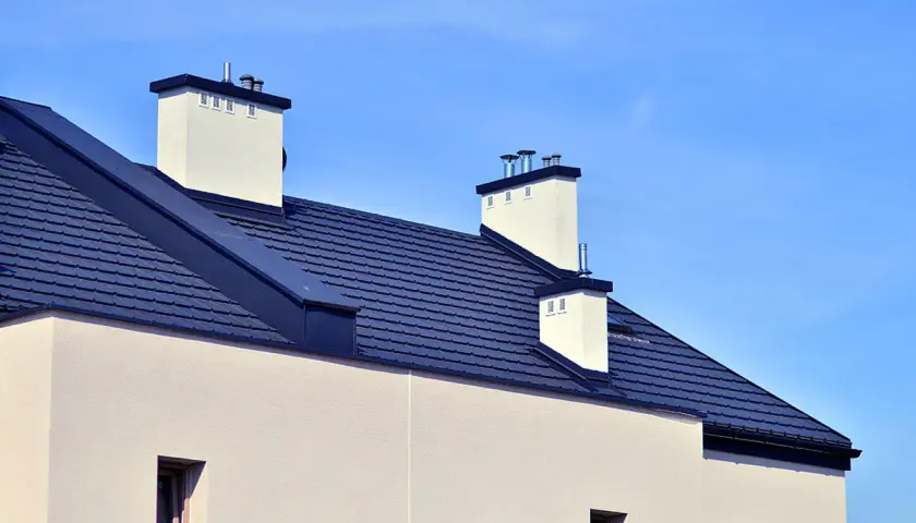Nowoczesny dach bezokapowy z ciemnogranatową dachówką i białymi kominami na tle błękitnego nieba.