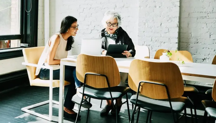 Zwei Frauen besprechen am Tisch die betriebsrente. Nachteile werden diskutiert, während sie auf Laptops und Tablets schauen.