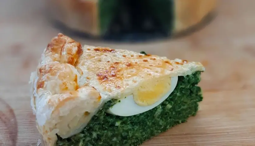 Kawałek torta pasqualina z zielonym nadzieniem i jajkiem, na drewnianej desce.