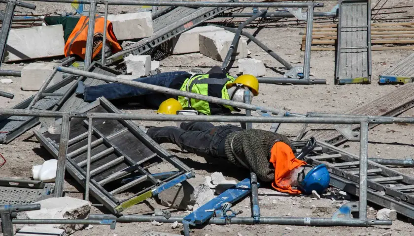 Dwa manekiny leżą na placu budowy wśród rusztowań i gruzu. Jeden ma żółty kask, drugi niebieski. Widać ich ubrania robocze i pomarańczowe kamizelki.