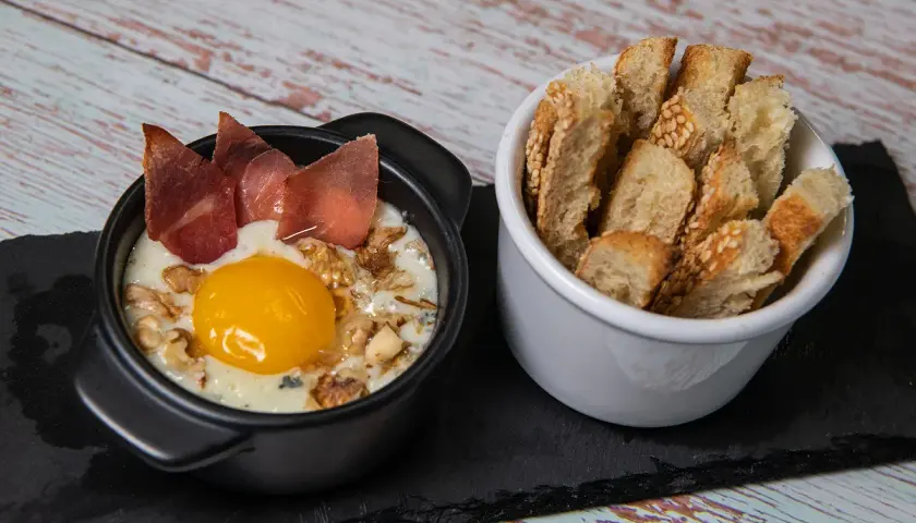 Un œuf cocotte raclette, crémeux et gourmand, garni de noix et de fines tranches de viande séchée, servi avec des toasts croustillants.