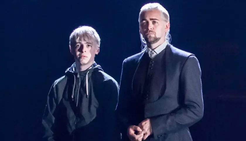 Scorpius Malfoy w czarnej bluzie obok starszego mężczyzny w eleganckim stroju.