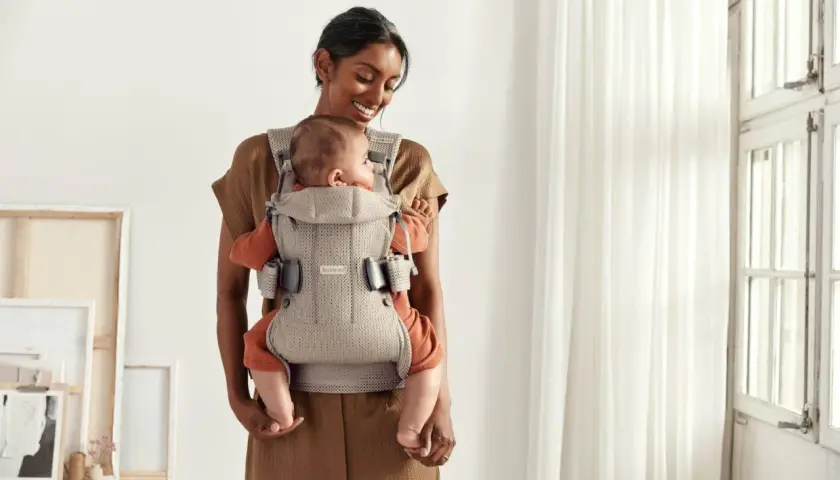 Uśmiechnięta mama tuli dziecko w nosidełku Babybjorn. Od kiedy można używać? Od pierwszych dni życia!