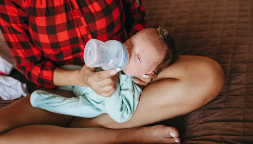 Mamma con maglia a quadri rossi offre il biberon a un neonato, praticando l'allattamento misto.