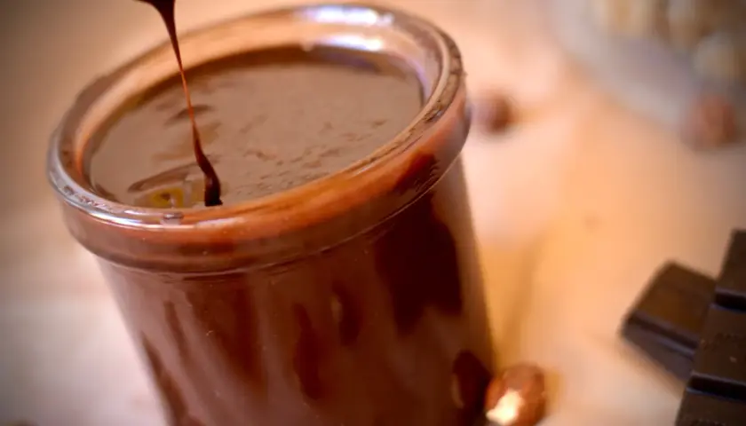 Un chorrito de crema de chocolate cae en un frasco lleno de esta delicia. Avellanas y tabletas de chocolate completan la escena.