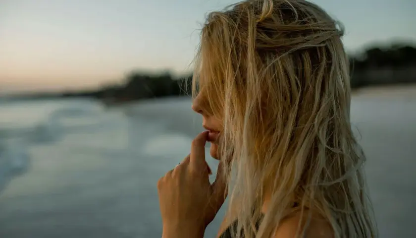 Kobieta z blond włosami rozjaśnionymi przy twarzy, na tle plaży i morza.