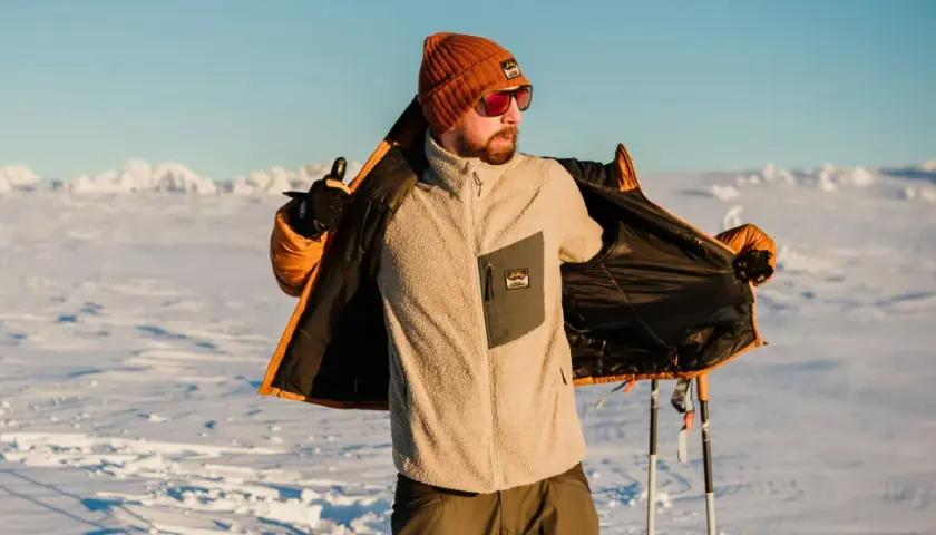 Man i orange mössa och solglasögon visar upp sin lager på lager-klädsel i snölandskap med skidstavar.