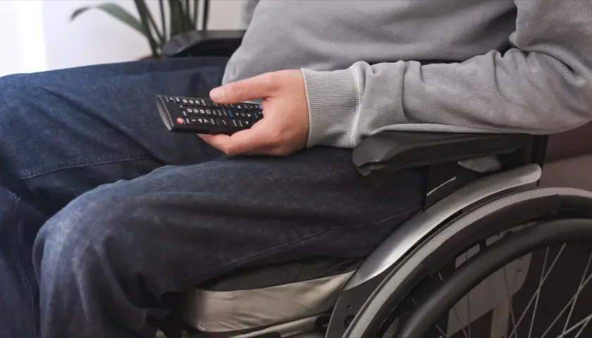 Osoba na wózku inwalidzkim, z umiarkowanym stopniem niepełnosprawności, trzyma pilot do telewizora.