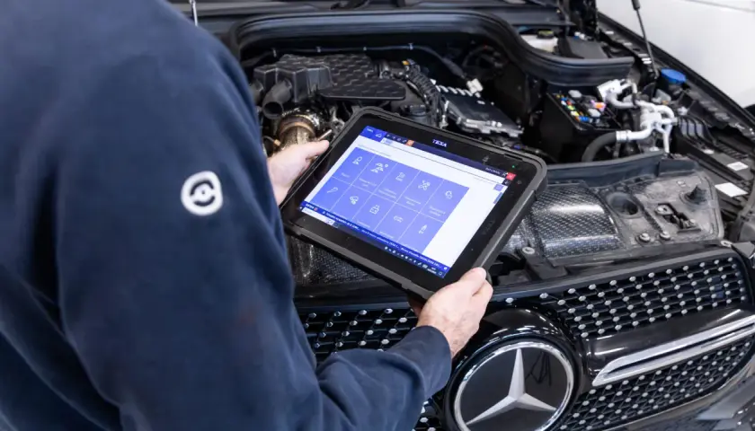 Meccanico esegue la prima revisione auto con tablet diagnostico davanti al motore di una Mercedes.