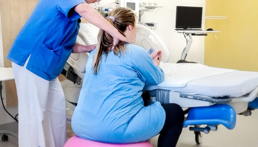 Barnmorska stöttar en kvinna på en rosa boll, v 34. De är i ett förlossningsrum med medicinsk utrustning.