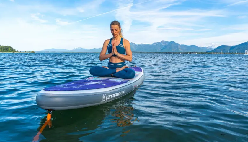 Frau praktiziert SUP Yoga auf einem Brett im See, mit Bergen im Hintergrund.