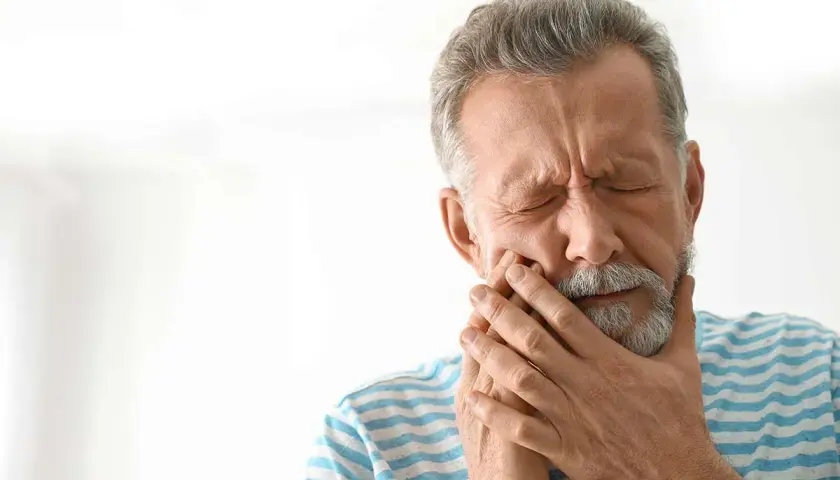 Hombre mayor con los ojos cerrados y la mano en la mejilla, sufriendo de dolor, posiblemente por alveolitis seca.