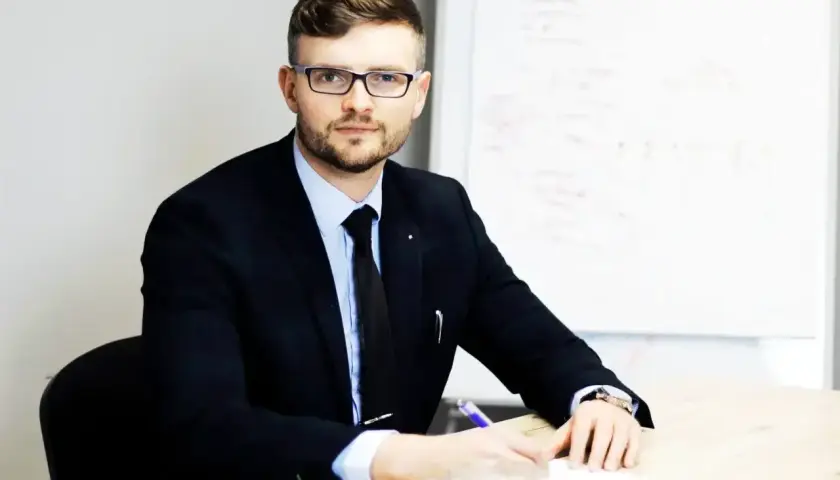 Mężczyzna w garniturze i okularach siedzi przy biurku, pisząc.