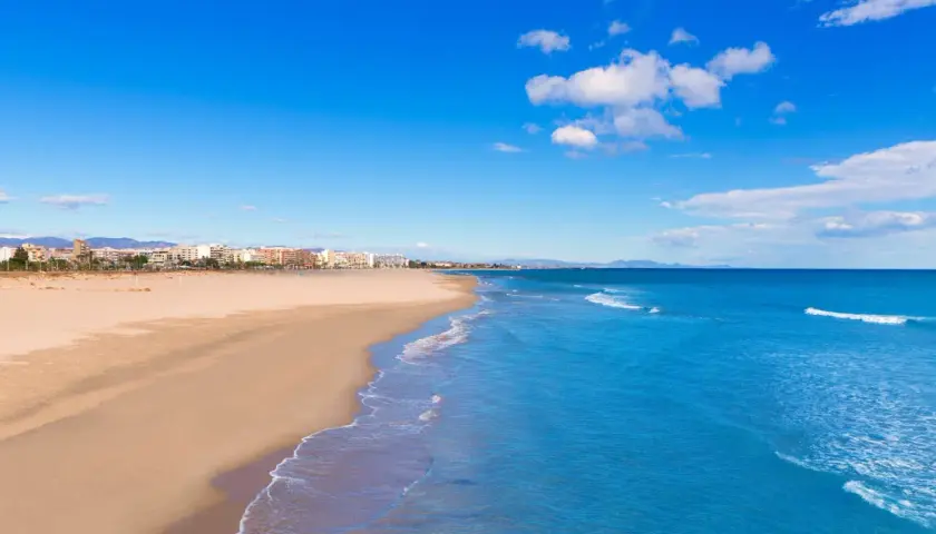 Amplia playa de arena dorada en Sagunto, con el mar Mediterráneo azul y edificios de la costa al fondo bajo un cielo despejado.