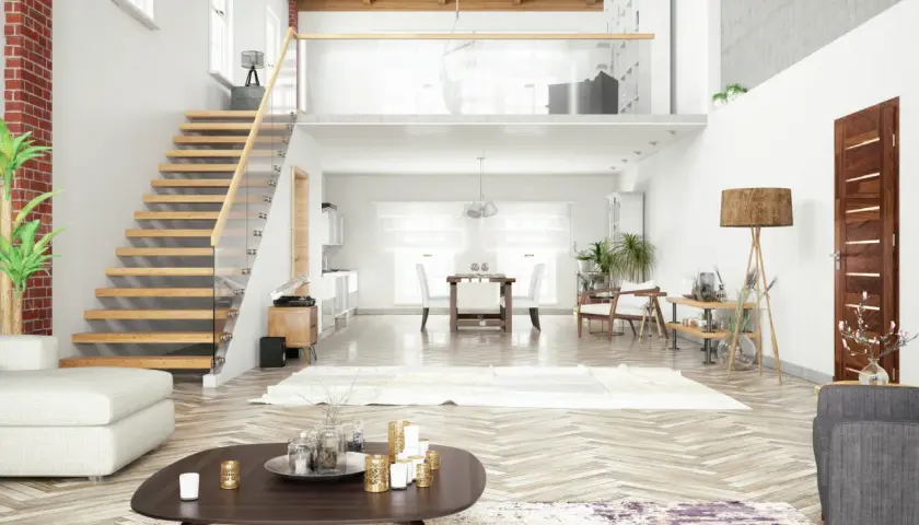 Un grand salon lumineux avec un escalier en bois menant à une mezzanine sous toit en pente. Un canapé blanc, une table basse et une salle à manger sont visibles.