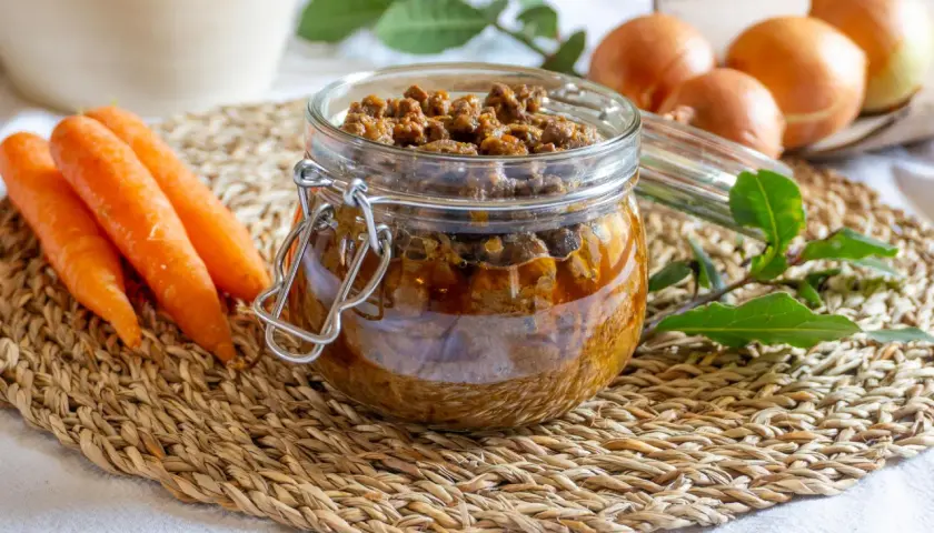 Un vasetto di vetro pieno di saporito ragù di anatra, con carote e cipolle sullo sfondo.