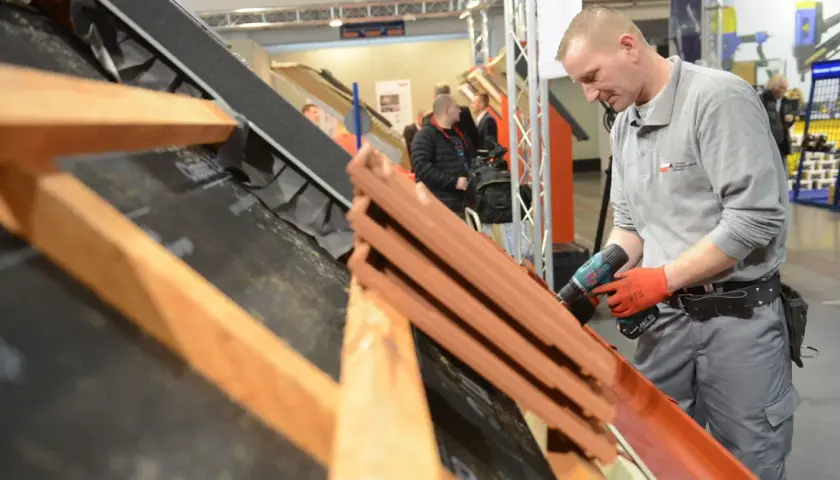 Dekarz w czerwonych rękawicach używa wkrętarki do montażu dachówek na konstrukcji dachu.